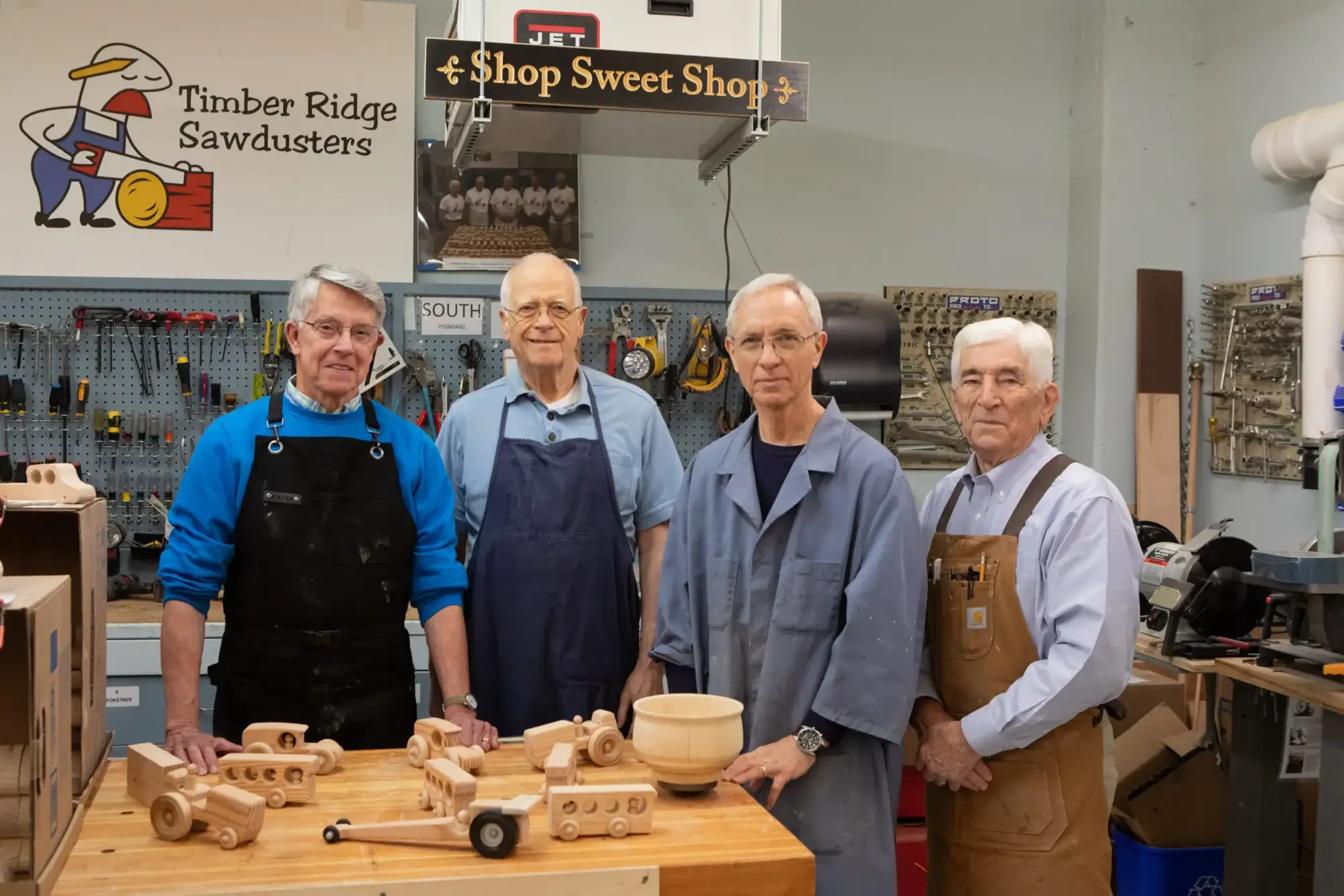 4 men standing behind a table filled with wood working tools, one of the many fun and engaging hobbies for seniors at Timber Ridge's wood shop.
