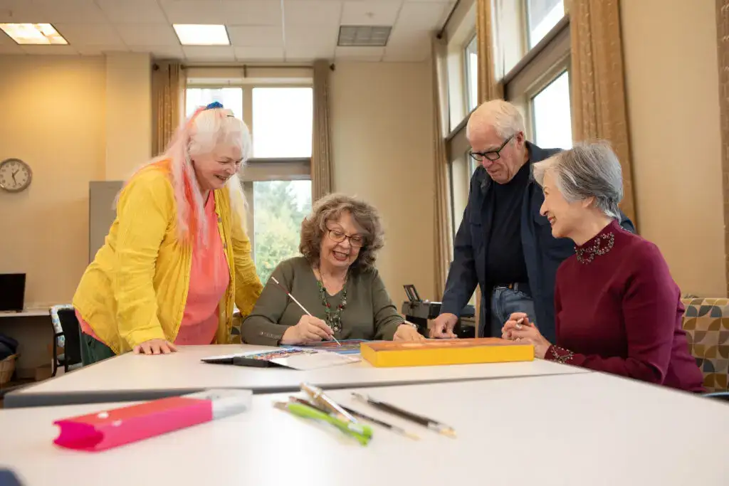 Residents of Timber Ridge at Talus in art studio, stand around table, watching a woman paint, one of the best hobbies for seniors in Issaquah, WA.