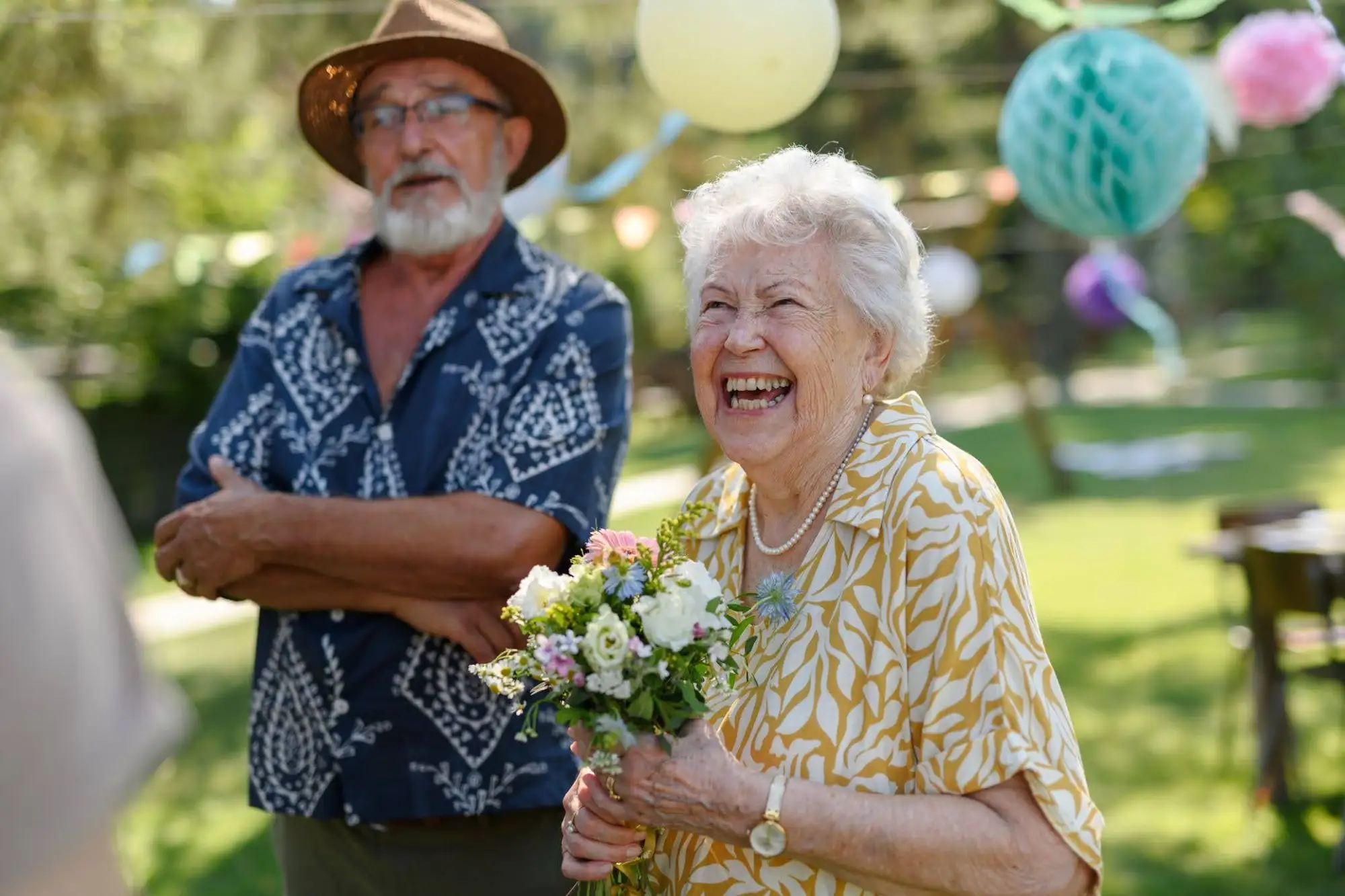 A senior woman holding flowers with her husband in the back at Timber Ridge at Talus, a senior living community that promotes modern retirement lifestyle in Issaquah, WA.